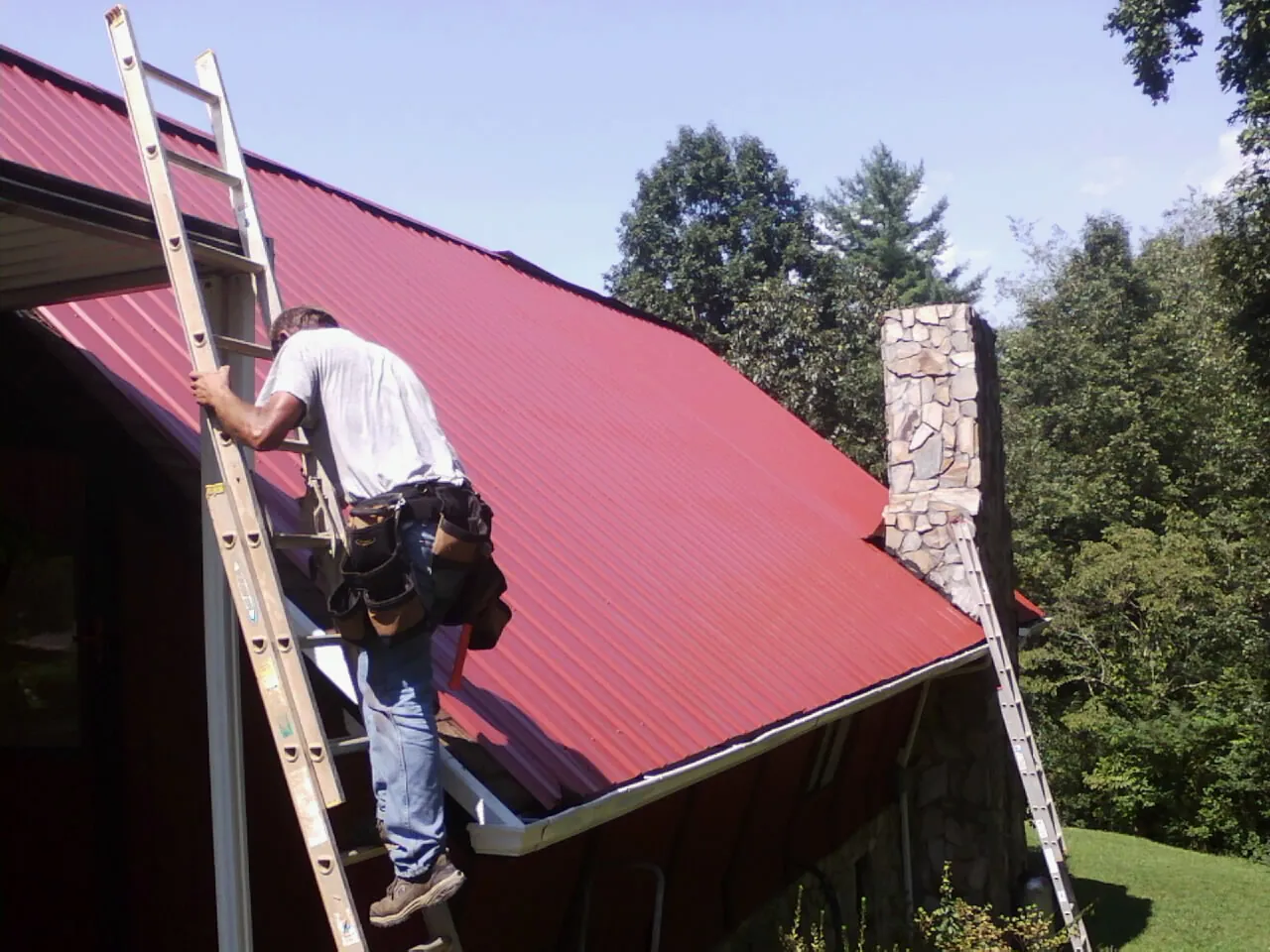 Roofer on a ladder beside a red metal roof and stone chimney — Soffit & Fascia Repair by Roofing Brothers in Newport News